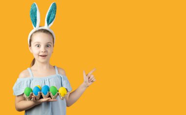 Mutlu Paskalyalar, çocuklar. Tavşan kulaklı bir kız elinde mavi ve sarı renkli yumurtalarla dolu bir tepsi tutuyor. Yüzünde şaşkınlık hissi var. İşaret parmağı boş bir alanı gösteriyor.