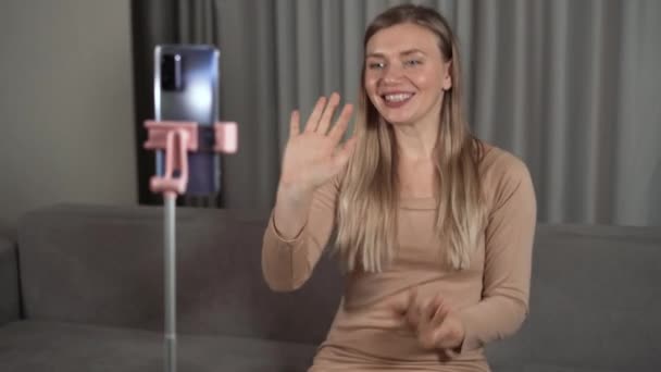 Portrait of a beautiful lady, a woman is sitting on the sofa and cheerfully waves her hand in front of a smartphone. concept of video call and community with loved ones. — Stock Video