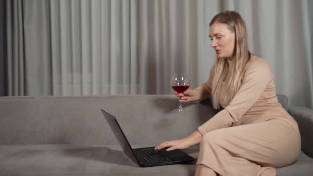 Glimlachende vrouw uit de 30 die een glas wijn drinkt terwijl ze thuis op de bank zit te chatten op een videoconferentie ter viering van een vakantiefeest in een virtuele chat, online ontmoeting met een laptop. — Stockvideo