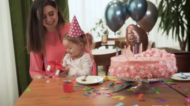 Mam en dochter zitten thuis aan tafel bij de taart en vieren hun verjaardag. Het meisje houdt een rode doos in haar handen.. — Stockvideo