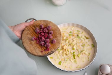 Masada hamurlu ve dilimlenmiş elmalı seramik bir kalıp var ve bir kadının elleri ahududu tutuyor. Evde, mutfakta meyveli turta yapma süreci. Ev yapımı pişirme..