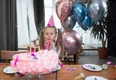 Küçük bir kız doğum gününü kutluyor, pastanın yanında tek başına duruyor ve neşeli bir pipo üflüyor. Hava balonları.