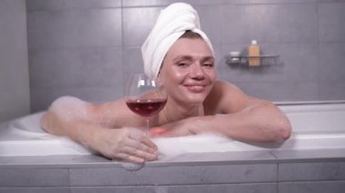 30 'lu yaşların mutlu genç kadını küvette oturmuş elinde şarap kadehiyle kameraya bakıyor. Mutlu bayan banyodan bir video görüşmesi yapıyor..