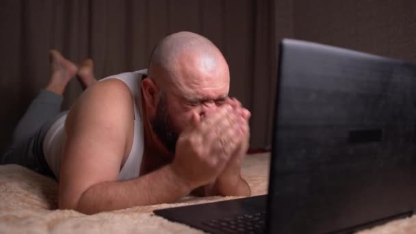 Un uomo barbuto adulto giace a casa su un letto vicino a un computer portatile, starnutisce e soffia il naso in un tovagliolo — Video Stock