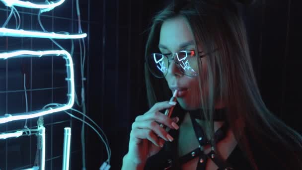 Mulher confiante e elegante com óculos em luz azul roxa fuma um cigarro eletrônico em uma festa perto de luzes de néon. Bonito feminino close-up — Vídeo de Stock
