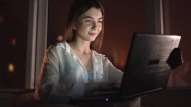 Genç, güzel, beyaz bir kadının yakın plan portresi büyük bir pencerenin yanında gece evde tek başına oturuyor. Gece hayatı. dizüstü bilgisayardaki klavyeye yazı yazmak. Boşluğu kopyala.