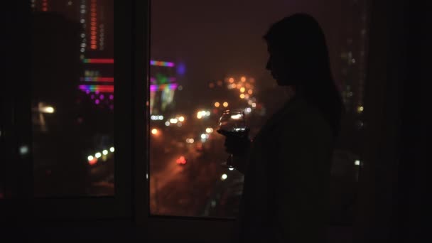 一个女人在夜晚凝视着城市，站在房子窗边的轮廓。拿着一杯葡萄酒和饮料。晚上一个人休息的概念. — 图库视频影像