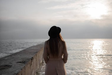 Açık renk montlu ve şapkalı genç bir kız rıhtımdaki deniz kıyısında duruyor ve uzaklığa bakıyor. İş kadını işten sonra su kenarında dinleniyor. Temiz hava bulutlu gökyüzü ve deniz.