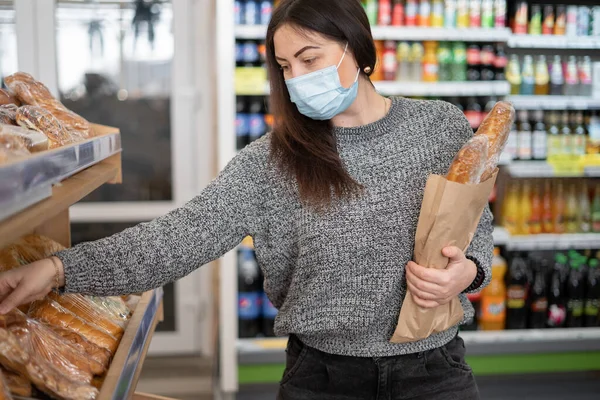 Young woman in a protective mask in the store buys fresh hot bread in the department. Shopping for food in quarantine.