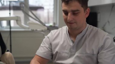 Yakışıklı esmer bir doktor. Dişçinin ofisinde oturuyor ve yüzüne tıbbi maske takıyor..