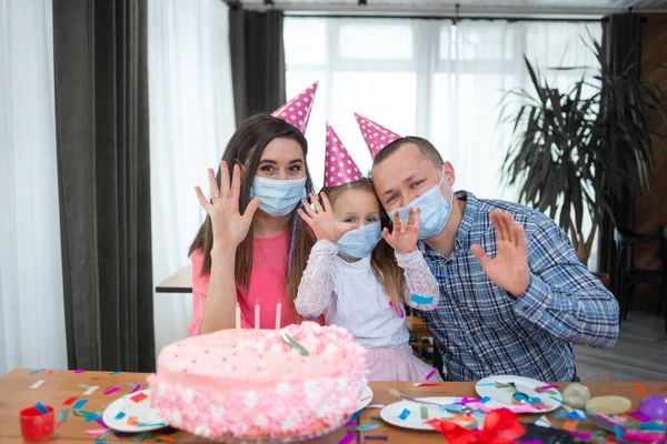 Caucasian family celebrating while sitting at home at the table. Medical masks and party caps, making a video call. birthday concept during a pandemic. Copy space.