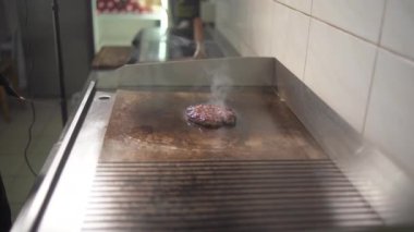 Peynirli burger için biftek ve domuz pirzolası pişiriyorum. Et mutfakta kızartılmış. Etrafta yağ damlaları ve duman var..