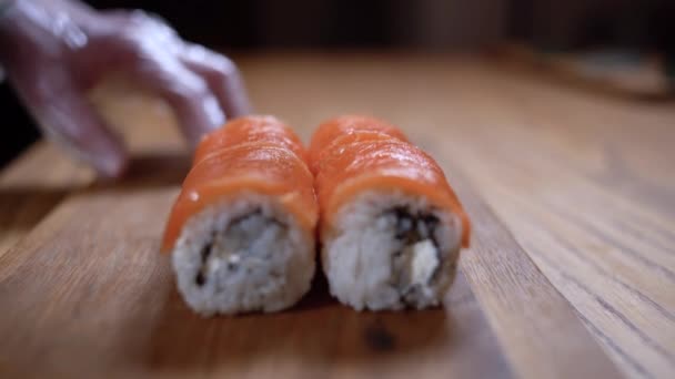 Les mains masculines coupent le rouleau de sushi de saumon en plusieurs morceaux sur une planche à découper. Coupe de rouleaux de sushi. — Video