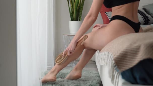 Eine Frau macht zu Hause eine Anti-Cellulite-Massage mit einer natürlichen trockenen Bürste. Sitzt auf dem Bett. Kopierraum. — Stockvideo