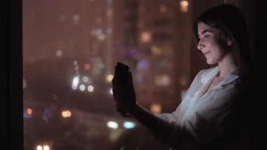 Kafkasyalı genç bir bayan gece pencerenin önünde dikiliyor, akıllı telefonuyla bir video görüşmesi yapıyor. Gece şehrinin ışıkları. Boşluğu kopyala.