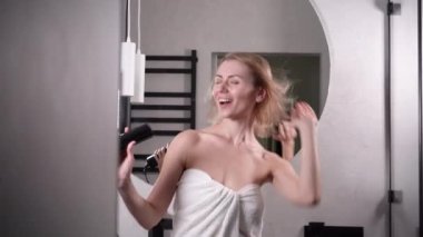 Genç, beyaz, güzel bir kadın saçını saç kurutma makinesiyle mutlu bir şekilde kurutuyor. Evde banyoda şarkılar söylüyor..