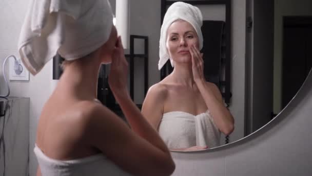 Szczęśliwa, atrakcyjna młoda kobieta z ręcznikiem na głowie, dotykająca twarzy, nakłada krem. spójrz w lustro, ciesz się naturalnym pięknem i zdrowym, czystym, nawilżonym środkiem do pielęgnacji skóry, zamknij — Wideo stockowe