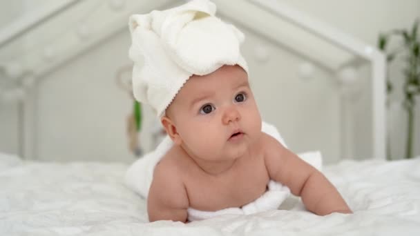 Ένα όμορφο νεογέννητο κοριτσάκι με μια πετσέτα στο κεφάλι ξαπλώνει σε ένα λευκό κρεβάτι, κοιτάζει τριγύρω και χαμογελάει. Έννοια ευτυχισμένων παιδιών. — Αρχείο Βίντεο