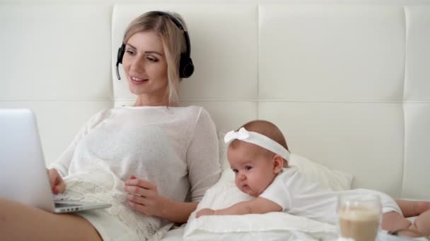 Joven madre sonriente con la niña usando el ordenador portátil mientras descansa en la cama en casa. Conexión a Internet. Red social. Videollamada en casa en la cama — Vídeo de stock