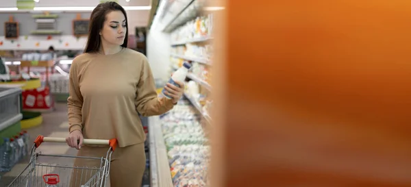 Happy young woman buys groceries at a grocery store. happy lady stands near the dairy display case and chooses yogurt. The buyer buys food at the market.
