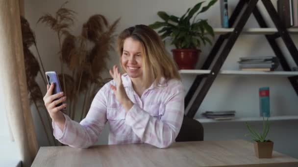 Unga vackra kaukasiska damen sitter vid bordet hemma. Gör ett videosamtal via programmet från sin smartphone. Plows hej med handen. — Stockvideo
