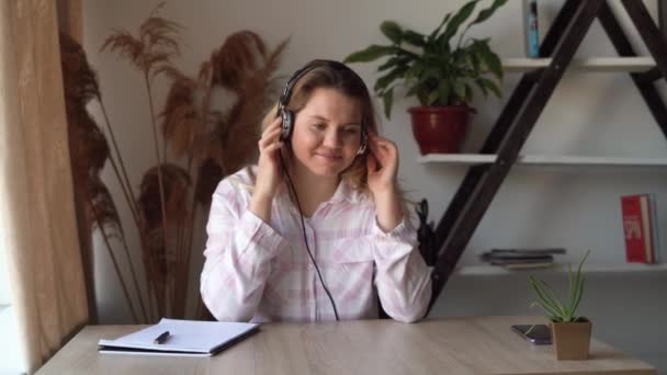 Jonge mooie blanke vrouw zit thuis aan een bureau, luistert naar muziek op koptelefoon en dansen. Favoriete melodie concept. — Stockvideo
