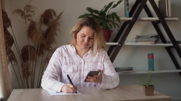 Een jonge mooie blanke dame zit thuis aan tafel, houdt haar smartphone in haar handen en maakt aantekeningen met een pen op een vel papier. De dag aan het plannen. Online opleiding. — Stockvideo