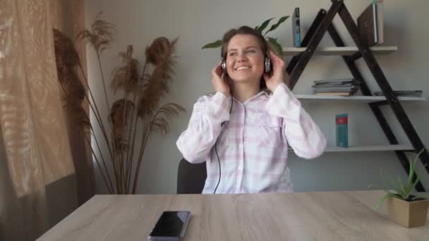 Jong mooi meisje voelt zich gelukkig, geniet van liedjes en dansen in het huis. Druk de headset tegen zijn oren en luistert naar muziek en dansen aan tafel. — Stockvideo