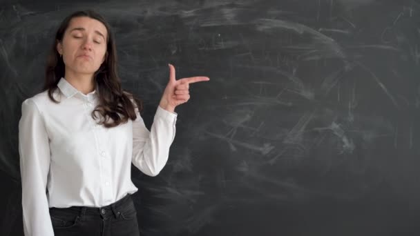 Studentessa caucasica si trova vicino alla lavagna di gesso, alza l'indice e indica uno spazio vuoto per il testo e annuisce affermativamente con la testa. copia spazio. — Video Stock