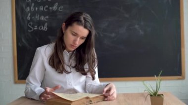 Genç güzel öğretmen tahtaya oturur ve bir kitap okur. Öğretmen masada oturuyor..