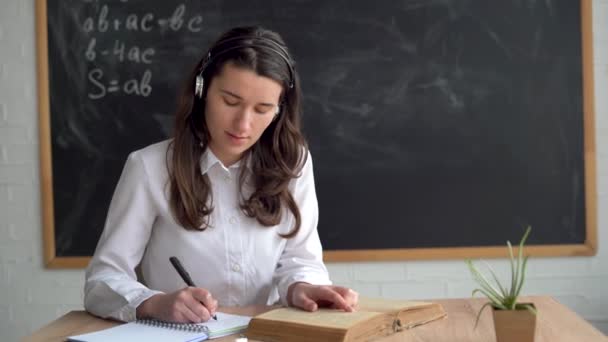 Joven hermosa maestra en auriculares con un micrófono se sienta delante de la pizarra y mira en un cuaderno. El profesor está sentado en la mesa. Lee un libro y toma notas en un cuaderno. — Vídeos de Stock