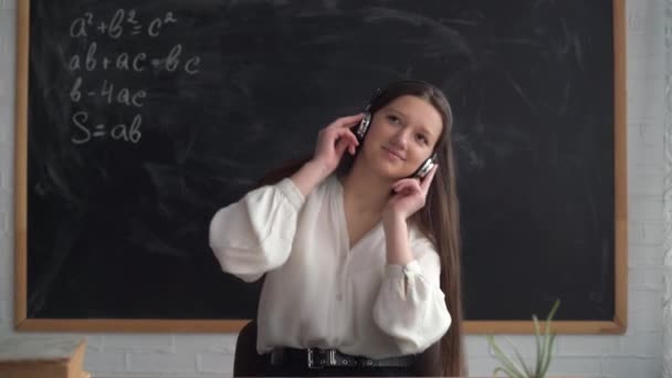 Girl student sits at the table, chalk board in the background. Headphones are wearing and the lady is dancing during the lesson. — Stock Video