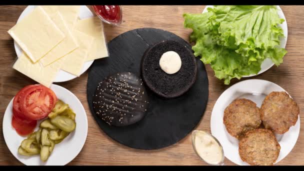 Hamburger ingredience, které tvoří lahodný burger jsou top view, zpomalený film, černá houska, salát a placka, sýr čedar na dřevěném stole. — Stock video