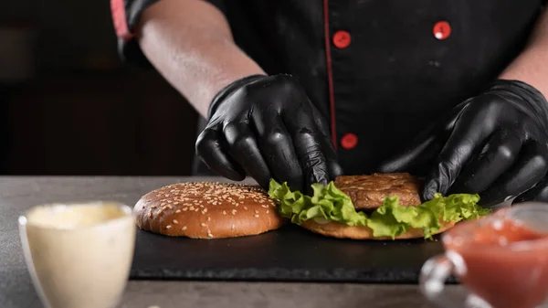 Receita passo a passo. Estágio 4. O chef prepara um delicioso hambúrguer. Close-up de uma mão em luvas pretas pondo uma costeleta em uma folha verde de alface, cardápio, comida deliciosa e insalubre. — Fotografia de Stock