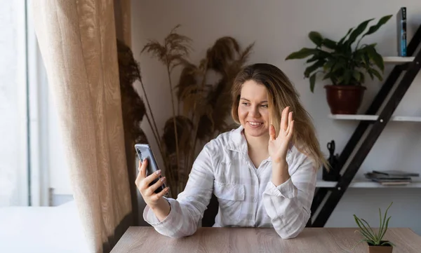 Портрет щасливої позитивної молодої жінки, яка спілкується через відеодзвінок у своєму смартфоні і сидить, посміхаючись, біля вікна. Дівчина в офісі з полицею з квітами і столом . — стокове фото