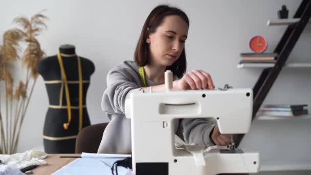 Die junge kaukasische Designerin arbeitet zu Hause im Büro. Eine Dame sitzt an einem Tisch und fädelt eine Nadel in einer Nähmaschine ein. Konzept für Kleinunternehmen. — Stockvideo