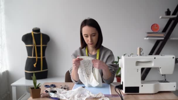 Een jonge mooie brunette zit aan een tafel in de buurt van een naaimachine, houdt damesslipjes in haar handen en controleert de kwaliteit van het naaien. Lady ontwerper naait lingerie. Kleine bedrijven. — Stockvideo