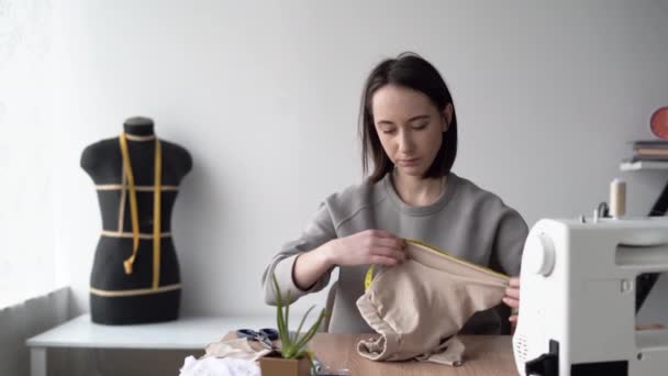 Jovem mulher caucasiana designer de moda está sentado em casa em seu escritório na mesa. Máquina de costura. Meça a peça de tecido necessária com um medidor flexível. Conceito de pequenas empresas. — Vídeo de Stock