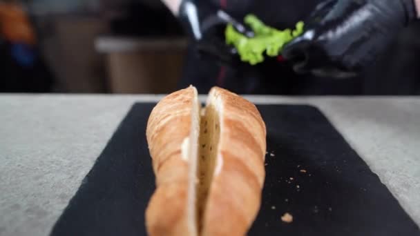 El primer plano de las manos del cocinero ponen la hoja verde de la lechuga sobre el croissant. El proceso de hacer un sándwich en la panadería. — Vídeo de stock