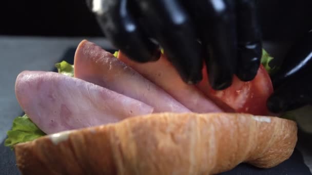 Primer plano de las manos de un chef con guantes negros preparando un sándwich de croissant. Desayuno rápido. Tomar rodajas de tomate y poner en el tocino. — Vídeos de Stock