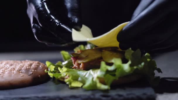 Proces výroby lahodného hamburgeru nebo cheeseburgeru. close-up Kuchaři ruku v rukavicích vloží kus sýra čedar do sendviče. — Stock video
