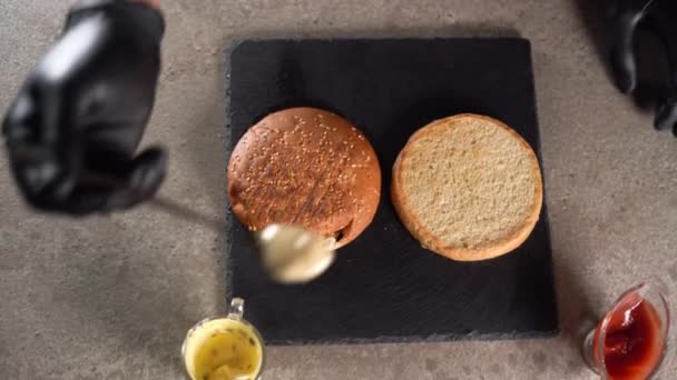 El proceso de hacer una deliciosa hamburguesa o hamburguesa con queso. close-up La mano enguantada de los chefs pone una cucharada de salsa blanca en el bollo. plano laico. — Vídeos de Stock