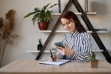 Genç, güzel, beyaz kız öğrenci evinde, masada oturuyor, akıllı telefonundan bir ders okuyor ve not defterindeki kalemle notlar alıyor..