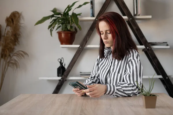 Молода красива білоруська бізнес-леді, сидячи за столом, тримає смартфон у руках, перевертаючи стрічку новин . — стокове фото