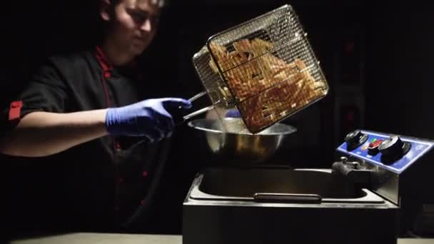 El chef prepara las papas fritas, saca una malla metálica de la freidora y vierte las papas fritas en un bol. — Vídeos de Stock