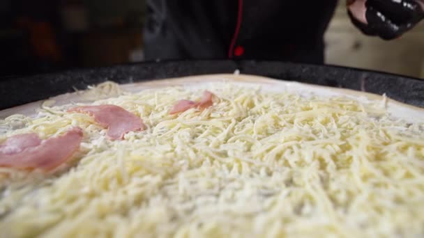 O processo de fazer uma pizza grande. As mãos do chef em luvas pretas põem o presunto no queijo mozzarella ralado. — Vídeo de Stock