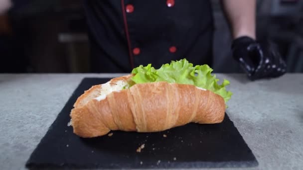 El primer plano de las manos del cocinero ponen la hoja verde de la lechuga sobre el croissant. El proceso de hacer un sándwich en la panadería. — Vídeo de stock