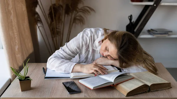 Πορτρέτο της καυκασίας νεαρή κουρασμένη κοπέλα που βρίσκεται σε ένα ξύλινο τραπέζι με βιβλία και εγχειρίδια στο σπίτι από το παράθυρο. Η γυναίκα ετοιμάζεται για τις εξετάσεις.. — Φωτογραφία Αρχείου