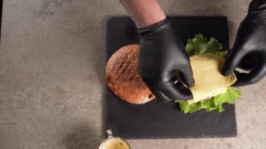Şef bir sandviç hazırlar. Şefin elinin yakın plan çekimi, lezzetli bir çift hamburger hazırlamak, sığır pirzolası üzerine peynir koymak ve üzerine susamlı çörek koymak. düz yat.