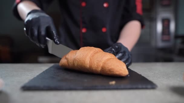 Las primeras manos de un chef con guantes cortan un croissant con un cuchillo afilado. Pasteles apetitosos en una tabla de cortar. Concepto de comida vegetariana saludable. — Vídeos de Stock